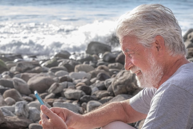 Ontspannen en lachende volwassen bebaarde senior man zittend op het rotsstrand met behulp van mobiele telefoon