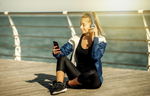 Ontspannen fitte vrouw in sportkleding met koptelefoon kiest muziek van een smartphone voor training op het strand op een zonnige dag