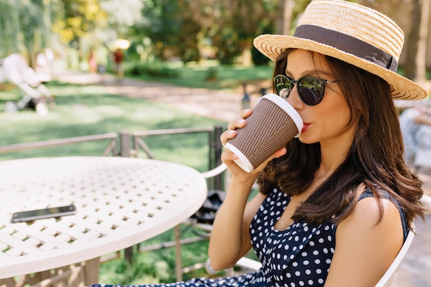 Ontspannen gelukkige vrouw in zonnebril en strooien hoed buiten koffie drinken