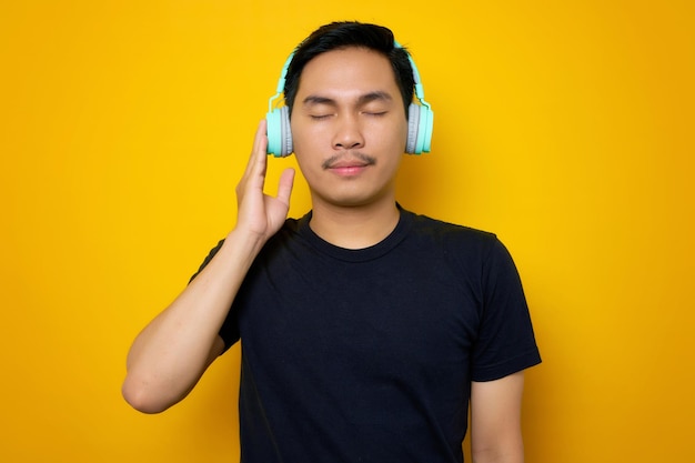 Ontspannen jonge Aziatische man in casual tshirt luisteren muziek in koptelefoon geïsoleerd op gele achtergrond Mensen emoties levensstijl concept