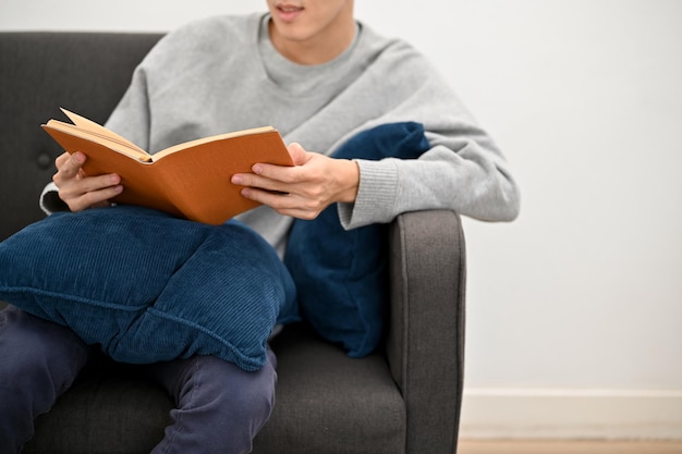 Ontspannen jonge Aziatische man zit op zijn bank in de woonkamer en leest graag een bijgesneden foto van een boek