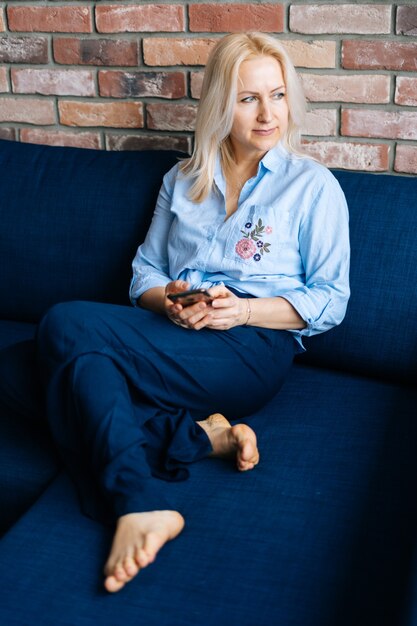 Ontspannen jonge blonde vrouw die mobiele telefoon gebruikt terwijl ze op een zachte donkerblauwe bank in de woonkamer zit