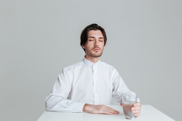 Ontspannen jonge man die waterglas vasthoudt terwijl hij aan de tafel zit geïsoleerd op de grijze muur