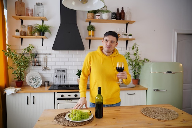 Ontspannen jonge man poseert met een glas rode wijn aan de keukentafel Volwassen man rust met alcohol en fruit in de keuken