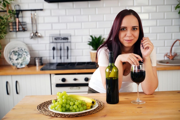 Ontspannen jonge vrouw drinkt rode wijn die aan de keukentafel staat Volwassen brunette rust met alcohol in de keuken