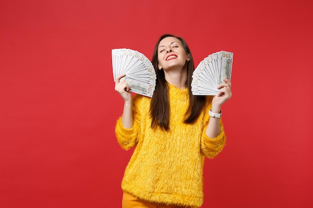 Ontspannen jonge vrouw in bonttrui met gesloten ogen met fan van geld in dollarbankbiljetten, contant geld geïsoleerd op rode achtergrond. Mensen oprechte emoties, lifestyle concept. Bespotten kopie ruimte.