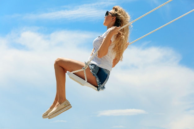 Ontspannen jonge vrouw met stijlvolle zomerkleren die tijdens warm weer hoog in de buurt van het meer zwaaien. Mooie blonde in zonnebril genieten van buitenactiviteiten.