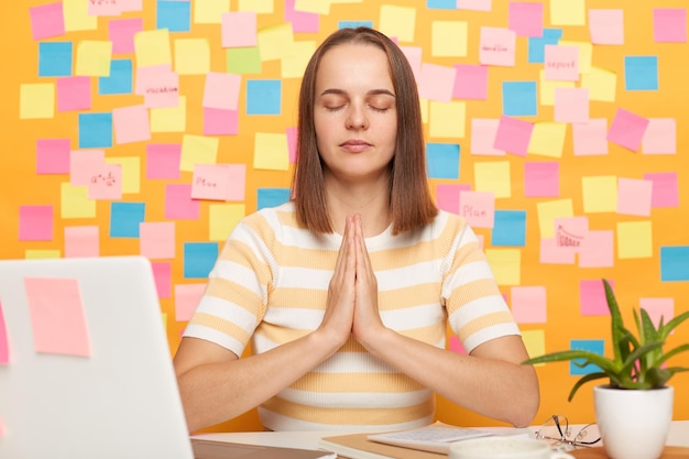 Foto ontspannen kalme vrouw in gestreept t-shirt zit op de werkplek op kantoor voor een notitieboekje tegen de gele muur met memokaarten houdt handen in biddend gebaar terwijl ze yoga beoefent terwijl ze pauze heeft op het werk