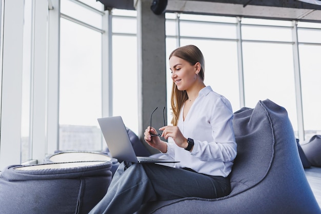 Ontspannen lachende gelukkige jonge vrouw in glazen en wit overhemd zittend in bean bag stoel rusten met behulp van laptop werk maken van aantekeningen in Kladblok computer remote werk concept