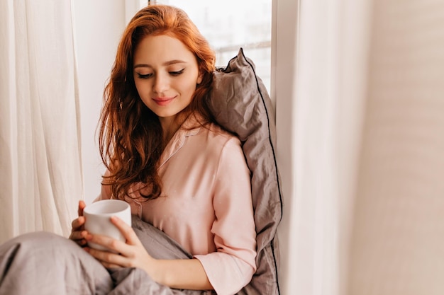 Ontspannen meisje in pyjama die koffie drinkt Binnenportret van knappe gembervrouw die van thee geniet