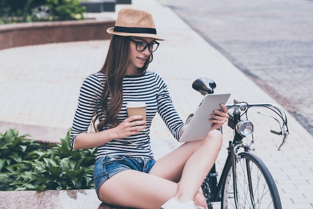 Ontspannen na de rit. Mooie jonge vrouw die naar haar digitale tablet kijkt en koffie drinkt terwijl ze op de bank bij haar fiets zit