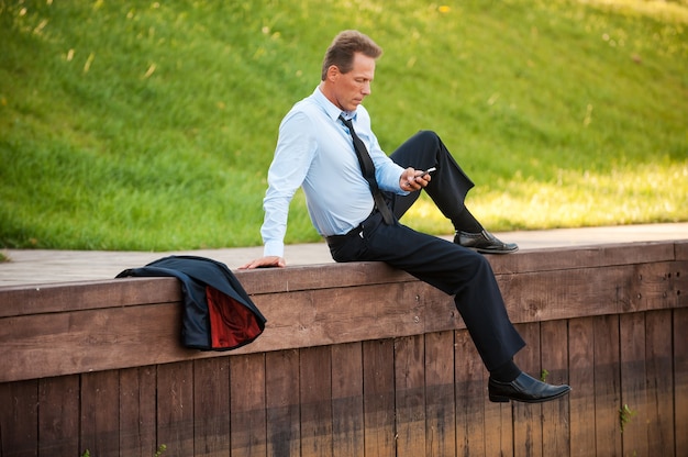 Ontspannen na een dag hard werken. Zelfverzekerde volwassen zakenman die mobiele telefoon vasthoudt en ernaar kijkt terwijl hij aan de kade zit