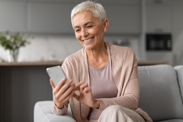 Ontspannen senior vrouw die graag mobiele apps op de telefoon binnen gebruikt