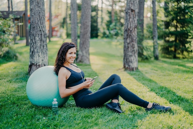 Ontspannen sportief jong vrouwelijk model neemt pauze na aerobicsoefeningen met fitnessbal zit op groen gras maakt gebruik van mobiele telefoonchats online luistert muziek in oortelefoons heeft atletisch lichaam