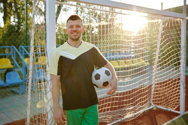 Foto ontspannen voetballer met bal staand leunt op voetbalpoort op het veld buiten vermoeide sportman die zich gelukkig en tevreden voelt als hij naar de camera kijkt