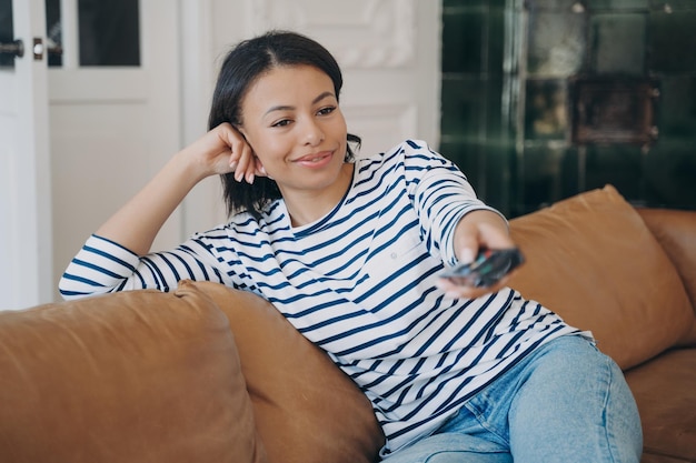 Ontspannen vrouw die televisie kijkt, verandert tv-zenders met de afstandsbediening die thuis op de bank zit