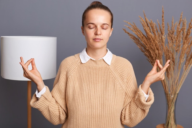 Foto ontspannen vrouw dragen beige trui poseren in kamer thuis staan met opgeheven armen en doen yoga mediteren oefening mudra gebaar poseren met gesloten ogen