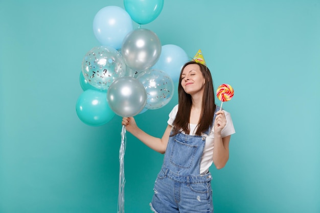 Ontspannen vrouw in verjaardagshoed die de ogen gesloten houdt en ronde lolly vasthoudt, vierend met kleurrijke luchtballonnen geïsoleerd op blauwe turkooizen achtergrond. Vakantie verjaardagsfeestje, mensen emoties concept.