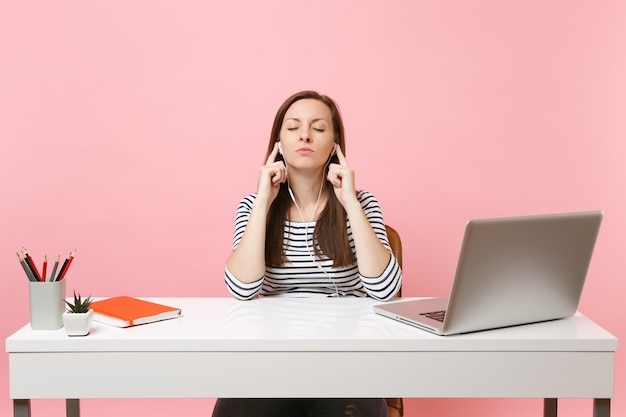 Ontspannen vrouw met gesloten ogen in oortelefoons luisteren muziek zitten en werken aan een wit bureau met hedendaagse pc-laptop geïsoleerd op pastel roze achtergrond. prestatie zakelijke carrière concept. ruimte kopiëren.