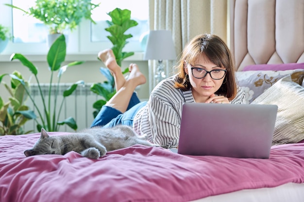 Ontspannen vrouw van middelbare leeftijd samen met slapende spinnende kat liggend op bed thuis vrouw met laptop voor vrije tijd werk rust Thuis vrije tijd levensstijl huisdier huisdier 40s mensen concept
