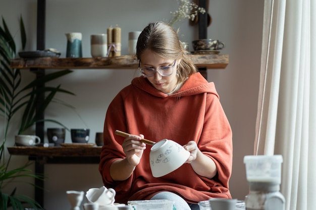 Foto ontspannen vrouwelijke artiest zit in een creatieve studio en tekent op handgemaakt porseleinen servies voor een ambachtelijke winkel