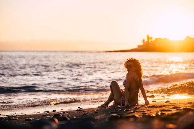 Ontspannen zittende mooie vrouw van middelbare leeftijd die naar de oceaan kijkt tijdens een gouden geweldige zonsondergang. gouden licht met oceaangolven vakantie en reislustconcept voor mensen