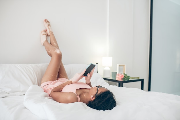 Ontspannend. Een vrouw in lingerie ligt in bed met haar benen omhoog en kijkt naar iets op een smartphone