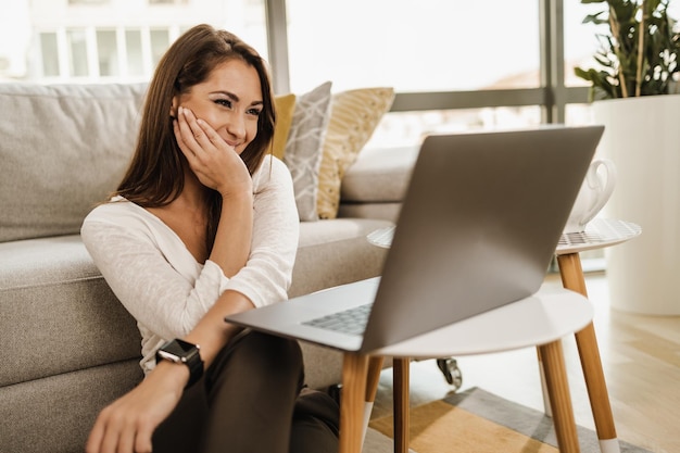 Ontspannende jonge vrouw zittend op de vloer in haar woonkamer en videogesprek voeren op haar laptop. Ze heeft vrije tijd op sociale media.