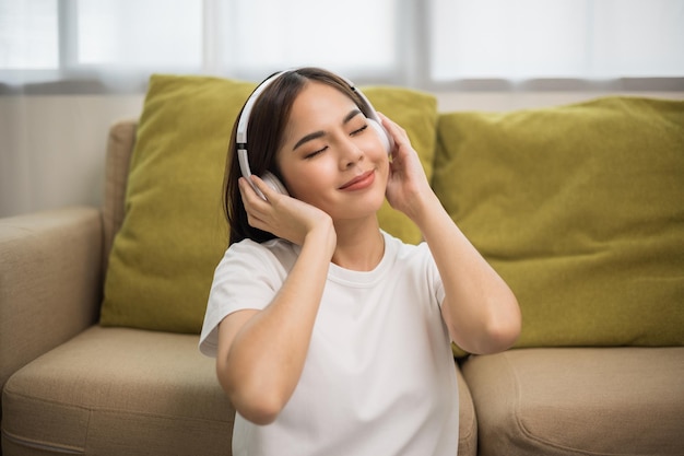 Ontspannende tijd op de bank. Mooie jonge aziatische vrouw die in de buurt van de bank zit, kiest muziek, rust uit en luistert naar een liedje met een koptelefoon. Lifestyle in de woonkamer thuis in de ochtend.