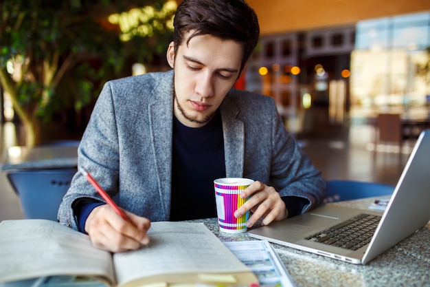 Ontsproten van jonge mannelijke studentenzitting bij lijst en het schrijven op notitieboekje. Jonge mannelijke student die in koffie bestudeert.