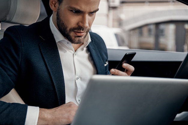 Ontwikkelen van nieuw project. Zelfverzekerde jonge man in volledig pak aan het werk met laptop terwijl hij in de auto zit