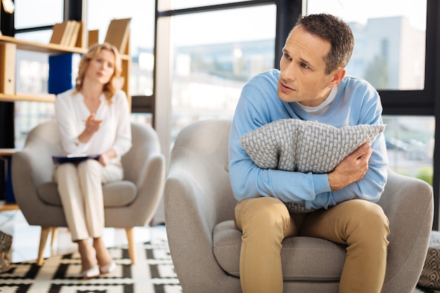 Onveilig voelen. Ongelukkige ongezellige depressieve man die een kussen vasthoudt en het knuffelt terwijl hij zich onveilig voelt