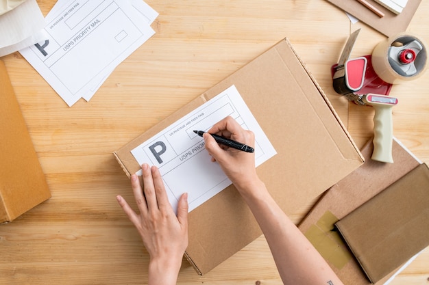 Photo onview of hands of young female manager of online shop holding black highlighter on noticepaper for client address on packed box