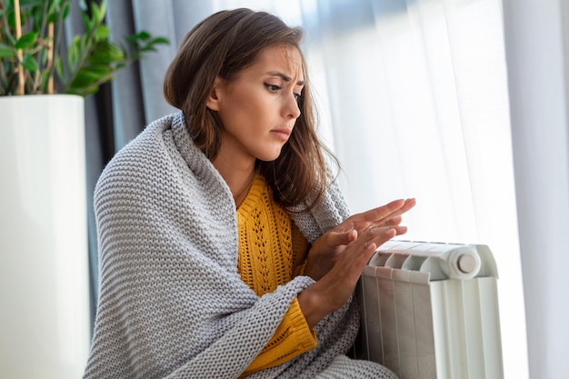 Onwel vrouw huurder in deken zit in koude woonkamer hand op oude radiator lijdt aan gebrek aan warmte Ongezonde jonge vrouw worstelt met koude bevriezing thuis Geen verwarmingsconcept