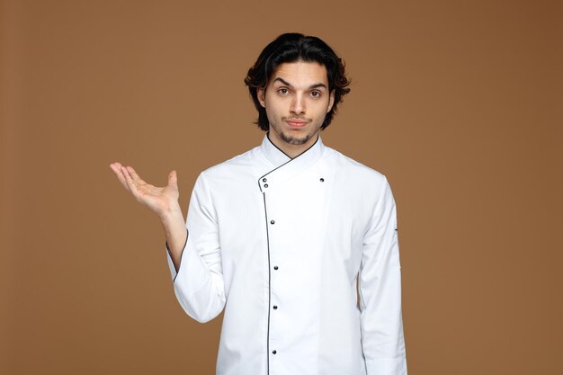 onzekere jonge mannelijke chef-kok in uniform kijkend naar camera met lege hand geïsoleerd op bruine achtergrond