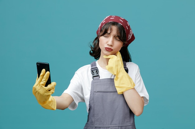 Onzekere jonge vrouwelijke schoonmaker draagt uniforme bandana en rubberen handschoenen die mobiele telefoon uitrekken en hand op de kin houden kijken naar mobiele telefoon denken geïsoleerd op blauwe achtergrond