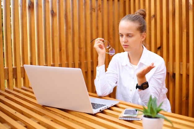 Onzekere, verbaasde vrouw, gekleed in een wit overhemd, zittend op een terras, werkend op een laptop, kijkend naar een computermonitor met een verbaasde uitdrukking