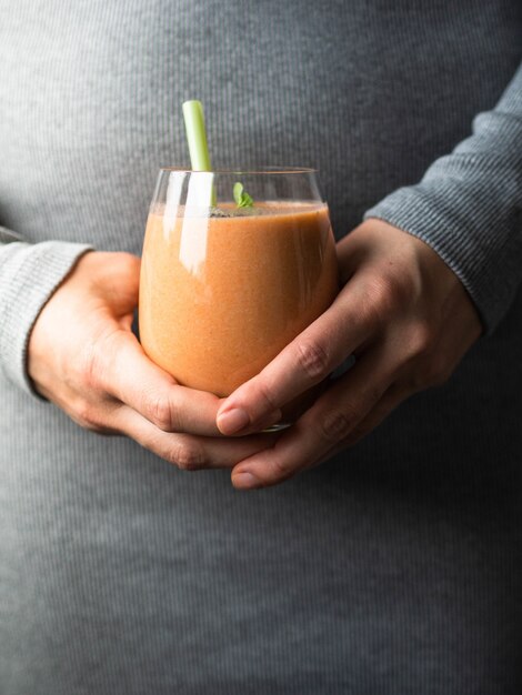 Onzekere vrouw die in grijze kleding een glas met oranje wortel smoothie houdt