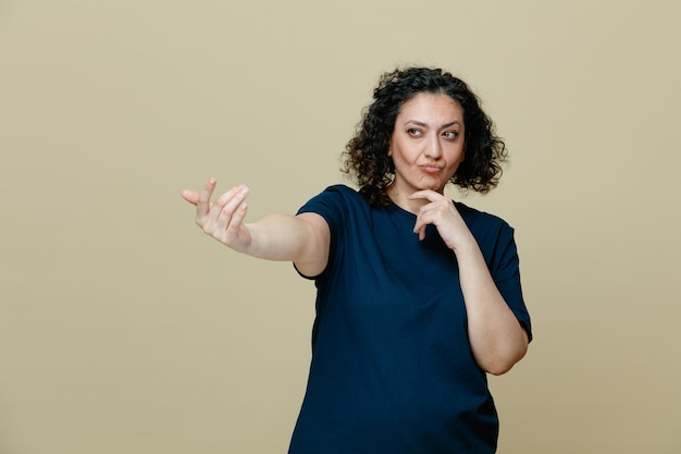 onzekere vrouw van middelbare leeftijd die een t-shirt draagt die de hand op de kin houdt en naar de zijkant kijkt en hier een gebaar maakt geïsoleerd op een olijfgroene achtergrond