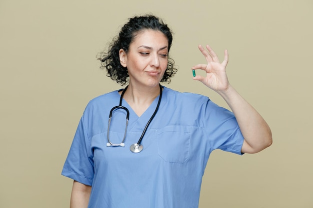Onzekere vrouwelijke arts van middelbare leeftijd die een uniform en een stethoscoop om de nek draagt en een capsule laat zien die ernaar kijkt geïsoleerd op een olijfachtergrond