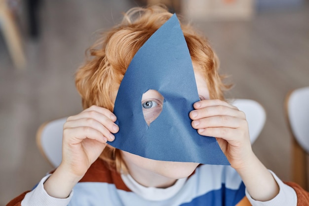 Oog van een schattige roodharige kleine leerling van de kleuterschool die naar de camera kijkt door een gat in donkere blu