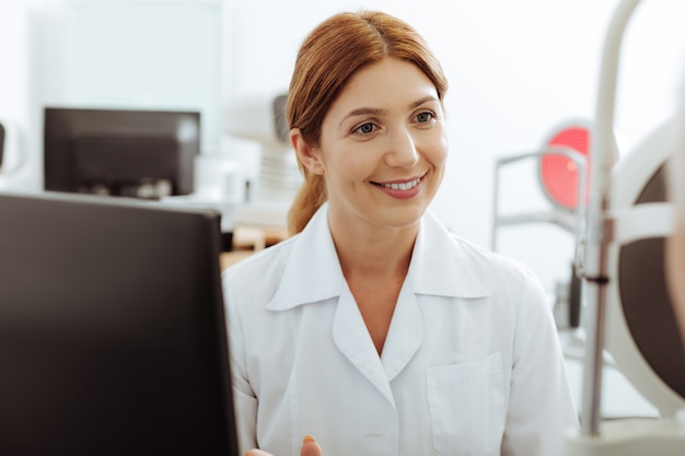 Oogspecialist zitten. Roodharige mooie gekwalificeerde oogspecialist zit aan de werktafel