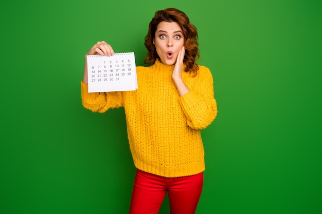 Ops! foto di donna piuttosto scioccata tenere il calendario cartaceo che mostra mese planner mano sulla guancia paura di essere incinta indossare maglione lavorato a maglia giallo pantaloni rossi isolati parete di colore verde