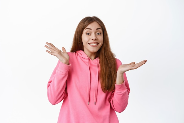 Oops sorry Smiling cute girl shrugging shoulders and apologizing for being unaware standing clueless dont know anything standing against white background