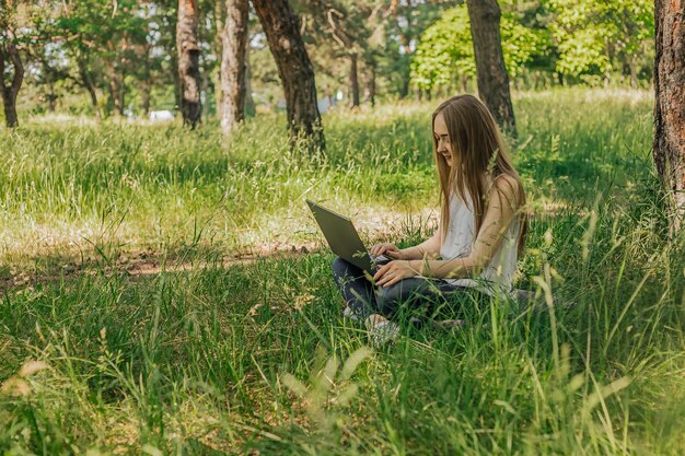 Op de banner werkt een jong meisje met een laptop in de frisse lucht in het park zittend op het gazon het concept van werken op afstand werk als freelancer het meisje volgt cursussen op een laptop en glimlacht