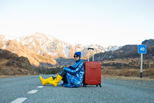 Op de bergweg zit een vrouw in een blauw jasje en hoed en knalgele laarzen met een rode koffer op de bergweggetjes