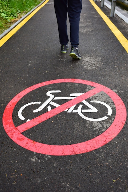 Op de geasfalteerde voetgangersstoep op de voorgrond staat een afdruk van een bord dat niet naar fietsers mag rijden. Op de achtergrond zijn de benen van een wandelende man in sneakers.