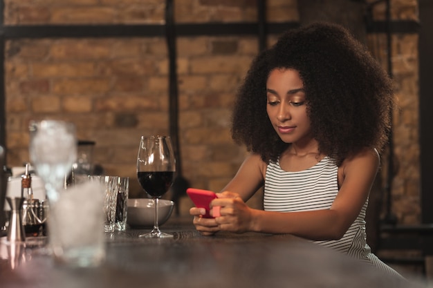 Op de hoogte blijven. Mooie krullende vrouw die haar telefoon gebruikt en op het web surft, recent nieuws leest, terwijl zij aan de bar zit en wijn drinkt