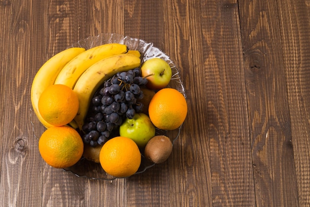 Op de houten tafel staat een schotel met verschillende soorten fruit Kiwi appels druiven bananen en sinaasappels