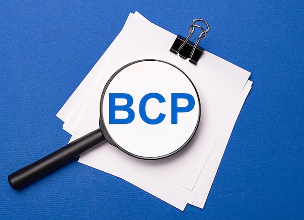 Op een blauwe achtergrond witte vellen onder een zwarte paperclip en daarop een vergrootglas met de tekst BCP Business Continuity Plan
